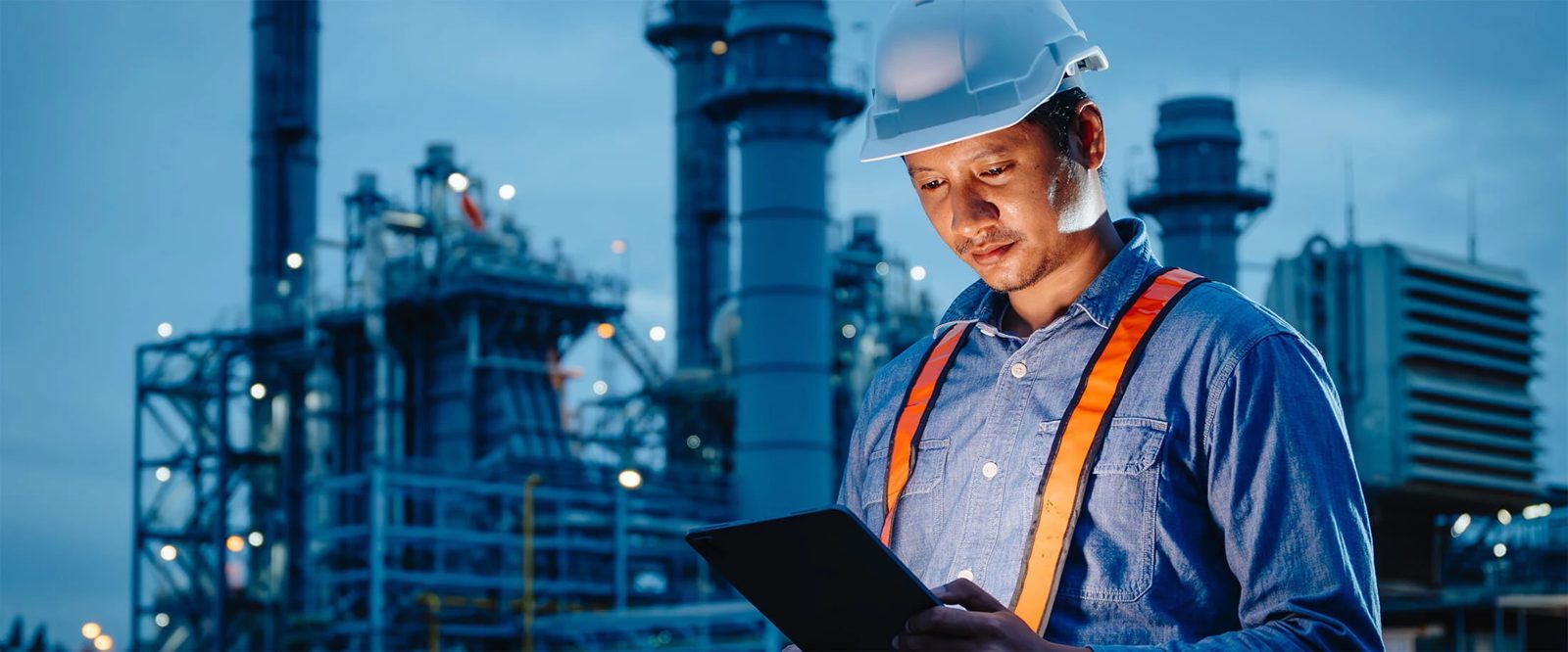 Technicien en casque de chantier blanc, chemise en jean et bretelles orange réfléchissantes, consultant une tablette tactile éclairée, devant un grand site industriel avec colonnes, passerelles métalliques et lumières, à la tombée de la nuit