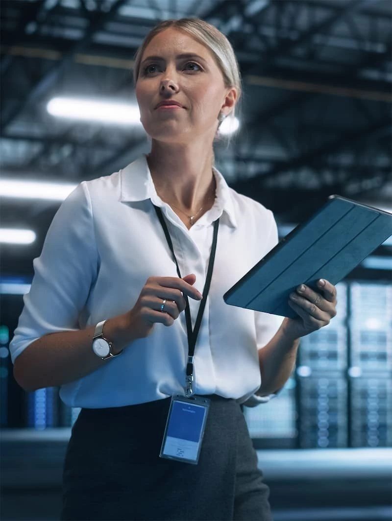 Conseillère en tenue professionnelle, chemisier blanc et badge autour du cou, tenant une tablette dans un local technique aux lumières bleutées, le regard tourné vers une installation