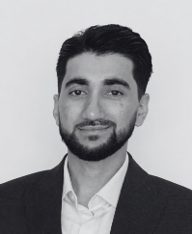 Portrait en noir et blanc de Mickaël, Conseiller en Radioprotection chez PCR Group : jeune homme aux cheveux bruns coiffés, barbe soignée, veste de costume sombre et chemise blanche, regard direct vers l’objectif