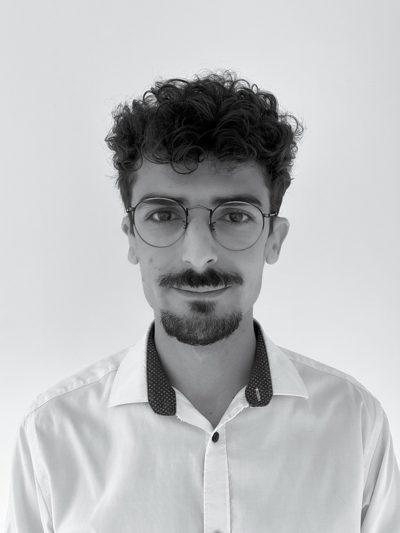 Portrait en noir et blanc de Victor Courtois, business developer de PCR Group : jeune homme aux cheveux bouclés, lunettes rondes, fine moustache et barbe, chemise blanche à col contrasté, souriant