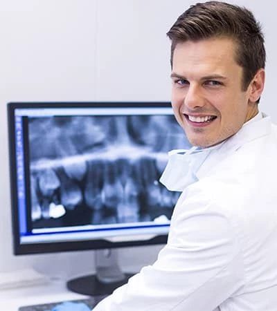 Chirurgien-dentiste souriant en blouse blanche, masque baissé autour du cou, assis devant un écran affichant une radiographie panoramique dentaire, dans un cabinet clair
