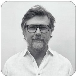 Portrait en noir et blanc de Cyril Jousset, fondateur de PCR Group et Conseiller en Radioprotection : homme aux cheveux grisonnants coiffés en arrière, lunettes à monture épaisse, barbe grise, chemise blanche, souriant légèrement face à l’objectif