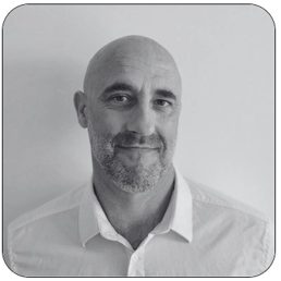 Portrait en noir et blanc de Lionnel Martin, Conseiller en Radioprotection chez PCR Group et ancien CRP en centrale nucléaire : homme au crâne rasé, barbe courte, chemise blanche à col ouvert, souriant devant un mur clair