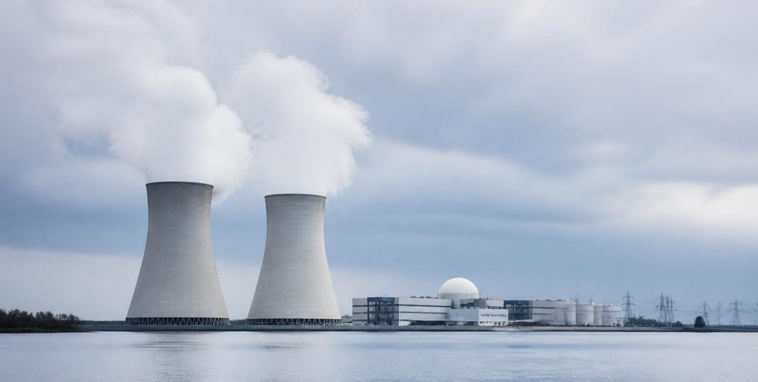 Deux tours de refroidissement d’une centrale nucléaire et le dôme blanc d’un bâtiment réacteur au bord d’un plan d’eau, sous un ciel nuageux gris, avec des panaches de vapeur